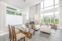A modern, bright living and dining area featuring large windows that allow natural light to fill the space. The dining area includes a glass table with wooden chairs, and a neutral-toned seating area with sofas and armchairs arranged around a minimalist fireplace. The decor is simple yet elegant, with a focus on clean lines and a contemporary aesthetic.