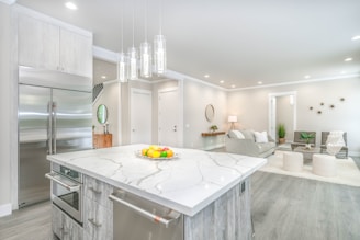 A modern, open-concept kitchen and living area with light gray tones. The kitchen features a large marble island with built-in appliances and a fruit bowl. Sleek, metallic refrigerator and pendant lights add a contemporary touch. The living area has neutral-colored sofas, a round mirror, and small decorative plants, with a floor lamp providing warm lighting.