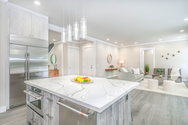 A modern, open-concept kitchen and living area with light gray tones. The kitchen features a large marble island with built-in appliances and a fruit bowl. Sleek, metallic refrigerator and pendant lights add a contemporary touch. The living area has neutral-colored sofas, a round mirror, and small decorative plants, with a floor lamp providing warm lighting.