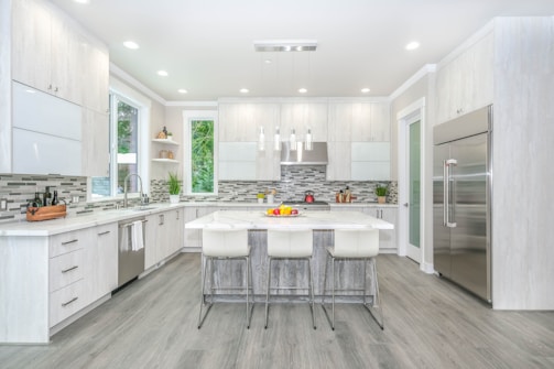 A modern kitchen with sleek white cabinetry and stainless steel appliances, featuring an island with bar stools in the center. The space is well-lit with ceiling lights and has large windows allowing natural light. The backsplash is a combination of gray and white tiles, providing a subtle contrast. Various kitchen items, including fruit bowls and kitchen utensils, decorate the countertops.