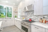 A modern kitchen with sleek white cabinetry and a light gray and white tiled backsplash. The countertops are a smooth, light-colored material, and a stainless steel oven and range hood are prominent. A red kettle sits on the stove, and decorative jars are placed on the counter. Natural light floods in from a large window, and green foliage is visible outside.