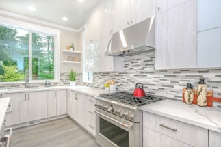 A modern kitchen with sleek white cabinetry and a light gray and white tiled backsplash. The countertops are a smooth, light-colored material, and a stainless steel oven and range hood are prominent. A red kettle sits on the stove, and decorative jars are placed on the counter. Natural light floods in from a large window, and green foliage is visible outside.
