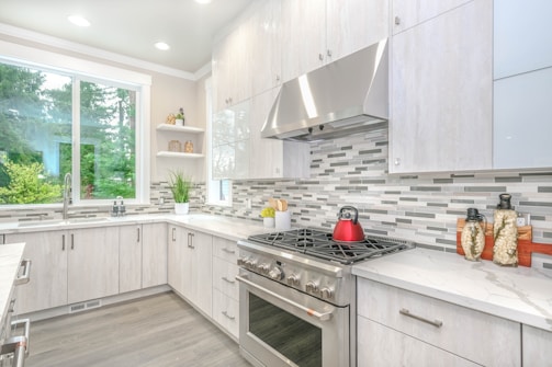 A modern kitchen with sleek white cabinetry and a light gray and white tiled backsplash. The countertops are a smooth, light-colored material, and a stainless steel oven and range hood are prominent. A red kettle sits on the stove, and decorative jars are placed on the counter. Natural light floods in from a large window, and green foliage is visible outside.