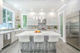 A modern kitchen with a large white marble island surrounded by four white stools. Stainless steel appliances, including a double-door refrigerator, add a sleek look. The backsplash features a contemporary gray and white tile design. Large windows provide natural light, enhancing the bright and clean aesthetic. Decorative plants and fruit bowls add a touch of color.