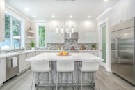 A modern kitchen with a large white marble island surrounded by four white stools. Stainless steel appliances, including a double-door refrigerator, add a sleek look. The backsplash features a contemporary gray and white tile design. Large windows provide natural light, enhancing the bright and clean aesthetic. Decorative plants and fruit bowls add a touch of color.