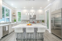 A modern kitchen with a large white marble island surrounded by four white stools. Stainless steel appliances, including a double-door refrigerator, add a sleek look. The backsplash features a contemporary gray and white tile design. Large windows provide natural light, enhancing the bright and clean aesthetic. Decorative plants and fruit bowls add a touch of color.