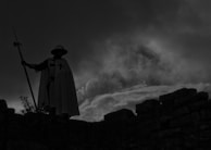 A silhouetted figure wearing a cloak and helmet stands atop a stone structure, holding a spear. Dark clouds form a dramatic backdrop, adding a sense of mystery and tension.