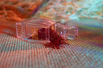 A clear plastic container lies on its side with its lid off, spilling a bunch of red, thread-like saffron strands onto a textured, woven surface. The background features a mix of blurred, colorful hues, including orange, green, and blue, creating a vibrant and warm ambiance.