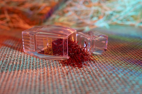 A clear plastic container lies on its side with its lid off, spilling a bunch of red, thread-like saffron strands onto a textured, woven surface. The background features a mix of blurred, colorful hues, including orange, green, and blue, creating a vibrant and warm ambiance.