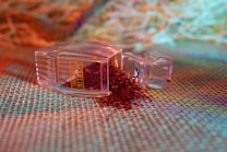A clear plastic container lies on its side with its lid off, spilling a bunch of red, thread-like saffron strands onto a textured, woven surface. The background features a mix of blurred, colorful hues, including orange, green, and blue, creating a vibrant and warm ambiance.