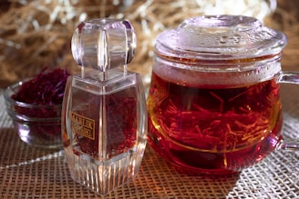 A steaming glass cup is filled with a rich, red liquid, likely tea, with saffron strands visible inside. Next to it, there is a small, transparent glass bowl containing vibrant red saffron threads. A large, clear, decorative container labeled 'Marlik Saffron' holds more saffron. The setting is on a wicker mat, with a blurred, warm-toned background.
