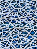 An intricate lattice pattern made of light-colored intersecting lines over a reflective glass surface. The lines create geometric shapes against a deep blue background, giving a sense of depth and complexity.