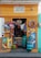 A small storefront with a vibrant yellow facade is adorned with various woven baskets, colorful ropes, and local advertisements for pet food and beverages. In the doorway, there are stacks of plastic containers and bunches of bananas.