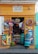 A small storefront with a vibrant yellow facade is adorned with various woven baskets, colorful ropes, and local advertisements for pet food and beverages. In the doorway, there are stacks of plastic containers and bunches of bananas.