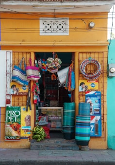 A small storefront with a vibrant yellow facade is adorned with various woven baskets, colorful ropes, and local advertisements for pet food and beverages. In the doorway, there are stacks of plastic containers and bunches of bananas.