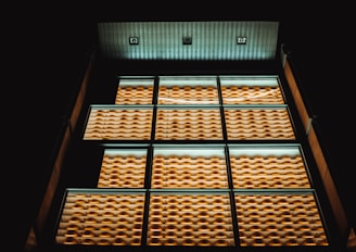 A modern building facade features rows of windows with uniform, bright yellow external features like louvers or shades. The lighting creates a warm glow against the dark background, highlighting the geometric pattern of the design.