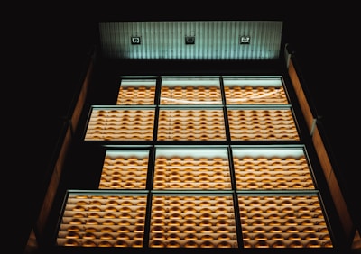 A modern building facade features rows of windows with uniform, bright yellow external features like louvers or shades. The lighting creates a warm glow against the dark background, highlighting the geometric pattern of the design.