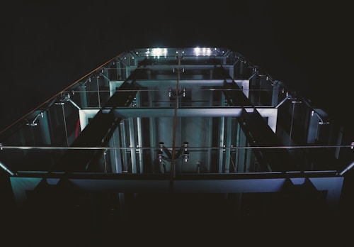 A tall structure with glass railings and visible metallic beams is illuminated by a series of lights, creating a modern and architectural appearance. The perspective gives a sense of looking up, highlighting the sleek design and symmetry of the structure against a dark background.