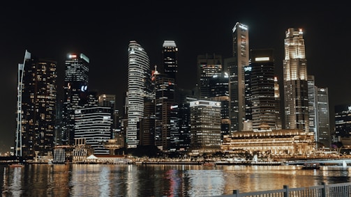 A sleek nighttime skyline of an international financial center with illuminated skyscrapers reflecting on water.