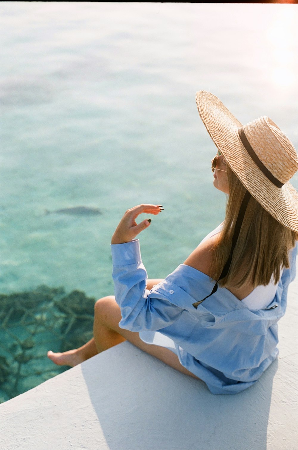 woman in white dress shirt and brown straw hat sitting on white sand beach during daytime