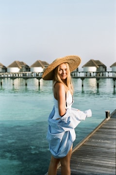 A woman stands on a boardwalk by the water, dressed in a white swimsuit and a large straw hat, holding a light blue garment. In the background, multiple overwater bungalows are visible, with serene, blue-green water underneath and clear sky above.