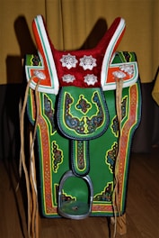 A colorful, ornate saddle with intricate patterns and decorations. It features a prominent red top with silver floral designs, green and orange accents, and detailed embroidery throughout. Leather straps hang on both sides, and there's a metallic stirrup at the bottom.