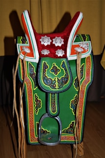 A colorful, ornate saddle with intricate patterns and decorations. It features a prominent red top with silver floral designs, green and orange accents, and detailed embroidery throughout. Leather straps hang on both sides, and there's a metallic stirrup at the bottom.