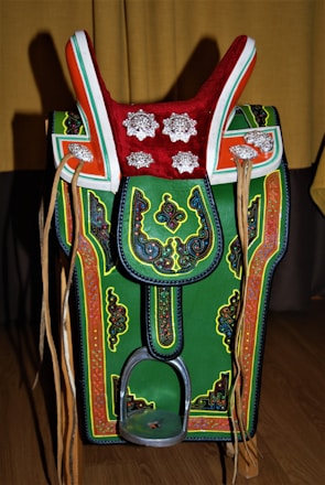 A colorful, ornate saddle with intricate patterns and decorations. It features a prominent red top with silver floral designs, green and orange accents, and detailed embroidery throughout. Leather straps hang on both sides, and there's a metallic stirrup at the bottom.