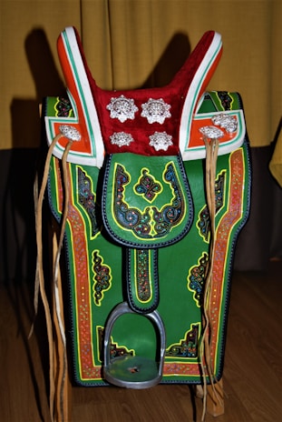 A colorful, ornate saddle with intricate patterns and decorations. It features a prominent red top with silver floral designs, green and orange accents, and detailed embroidery throughout. Leather straps hang on both sides, and there's a metallic stirrup at the bottom.