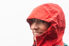 A person smiling while wearing a red hooded rain jacket with droplets of water visible on the fabric. The background is completely white, suggesting an overcast or foggy environment.