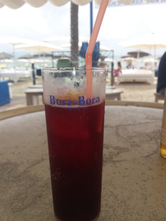 A tall glass filled with a dark red beverage and a pink straw, placed on a textured outdoor table. The glass has 'Bora-Bora Ibiza' written on it. In the background, there is a beach setting with blurred images of sun loungers, umbrellas, and people, creating a relaxed atmosphere.