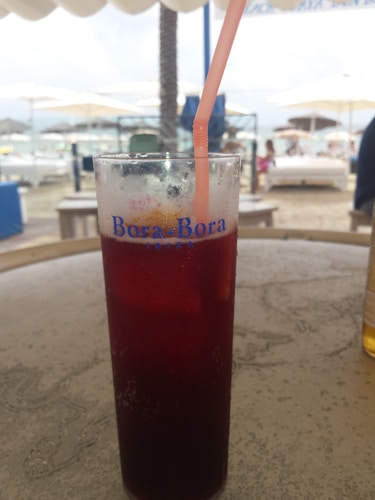 A tall glass filled with a dark red beverage and a pink straw, placed on a textured outdoor table. The glass has 'Bora-Bora Ibiza' written on it. In the background, there is a beach setting with blurred images of sun loungers, umbrellas, and people, creating a relaxed atmosphere.