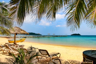 A tropical beach setting with white sand, clear blue waters, and gentle waves. There are palm trees providing shade, a series of lounge chairs positioned for relaxation, and a small yellow boat resting on the shore. In the distance, a wooden pier extends into the ocean, equipped with straw umbrellas for shade. An island covered with greenery can be seen on the horizon. The sky is partly cloudy with bright sunlight enhancing the vivid colors of the scene.