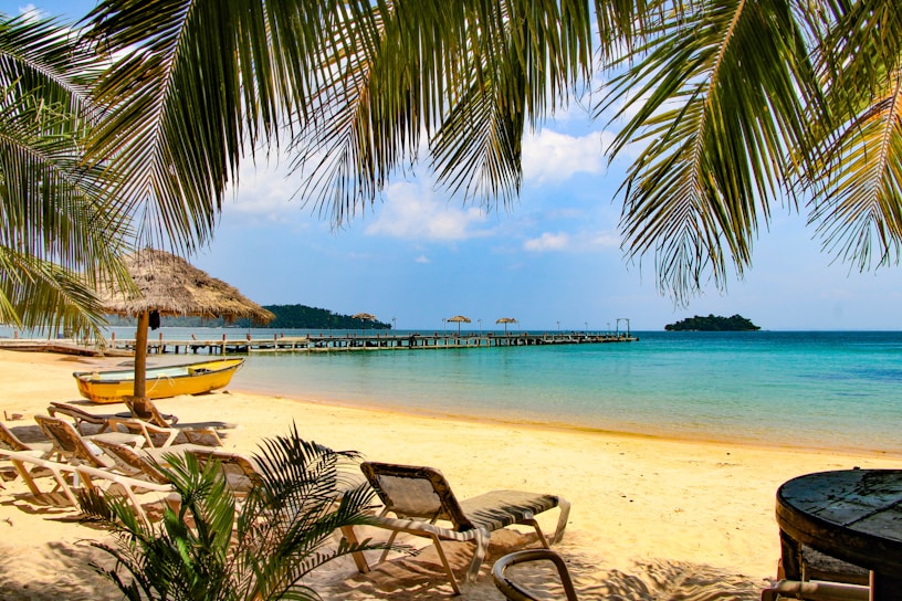 A tropical beach setting with white sand, clear blue waters, and gentle waves. There are palm trees providing shade, a series of lounge chairs positioned for relaxation, and a small yellow boat resting on the shore. In the distance, a wooden pier extends into the ocean, equipped with straw umbrellas for shade. An island covered with greenery can be seen on the horizon. The sky is partly cloudy with bright sunlight enhancing the vivid colors of the scene.