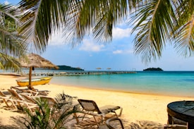 A tropical beach setting with white sand, clear blue waters, and gentle waves. There are palm trees providing shade, a series of lounge chairs positioned for relaxation, and a small yellow boat resting on the shore. In the distance, a wooden pier extends into the ocean, equipped with straw umbrellas for shade. An island covered with greenery can be seen on the horizon. The sky is partly cloudy with bright sunlight enhancing the vivid colors of the scene.