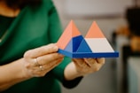 Hands are holding a geometric model consisting of colorful triangular pyramids placed on a blue square base. Each pyramid features different color combinations including red, blue, and white.