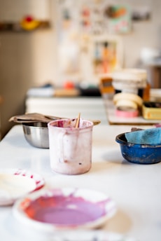 A workspace with painting supplies including bowls, containers with paint, and brushes. The background has blurred images and objects, suggesting an artistic environment.