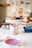 A workspace with painting supplies including bowls, containers with paint, and brushes. The background has blurred images and objects, suggesting an artistic environment.