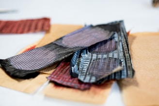 A collection of intricately patterned and textured paper strips lies on a flat surface. The pieces showcase geometric designs with a mix of colors, prominently featuring dark hues and some reddish tones. The papers appear to be layered in a casual arrangement.