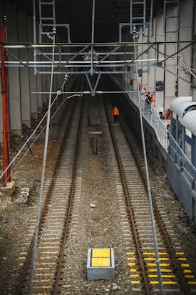 A railway track extends into the distance, flanked by tall walls and overhead wiring which are part of the infrastructure. Several workers in orange safety vests are seen along the right side walkway, with one standing on the track. The scene is enclosed and industrial, with cables and support structures visible above.