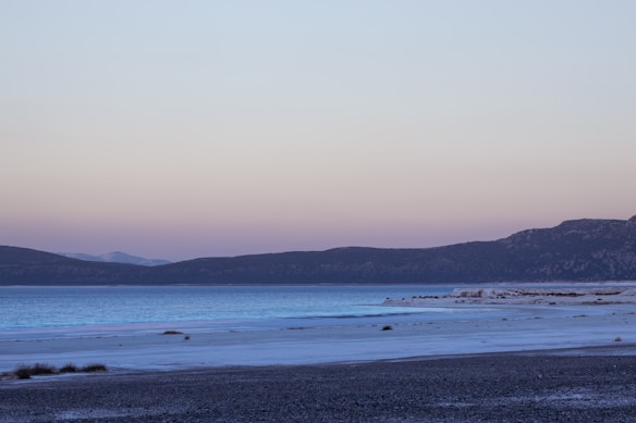 A serene lakeside scene with clear, still water reflecting the soft hues of a setting or rising sun. The sky is a gradient of pastel pink and blue, with distant hills providing a gentle backdrop. Sparse vegetation and patches of sand are visible along the shore.