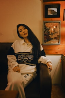 A person with long dark hair is seated on a leather couch in a warmly lit room with wooden accents. They are wearing a white and black sweatshirt with the word 'Explorer' and other text printed on it, along with white pants. The background wall features framed pictures, one of which is an old magazine cover from 'Popular Mechanics'.