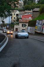 A small convertible car is driving on a curving paved road in a suburban area. Other cars are visible in the background, with their headlights on. The road is lined with houses, greenery, and several billboards. A traffic light is visible, showing green, and the area appears to be slightly overcast.