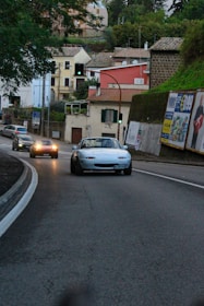 A small convertible car is driving on a curving paved road in a suburban area. Other cars are visible in the background, with their headlights on. The road is lined with houses, greenery, and several billboards. A traffic light is visible, showing green, and the area appears to be slightly overcast.