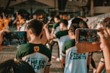 People wearing matching 'Cheer Cadets' uniforms participate in a group activity in an outdoor area, while two individuals hold up their smartphones to capture the moment. In the background, other groups in different colored uniforms are visible, along with drums and other equipment.