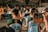 People wearing matching 'Cheer Cadets' uniforms participate in a group activity in an outdoor area, while two individuals hold up their smartphones to capture the moment. In the background, other groups in different colored uniforms are visible, along with drums and other equipment.
