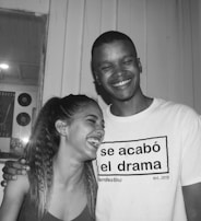 A black and white photograph of two people laughing, standing closely together. They appear to be enjoying a lighthearted moment. One person is wearing a T-shirt with Spanish text.