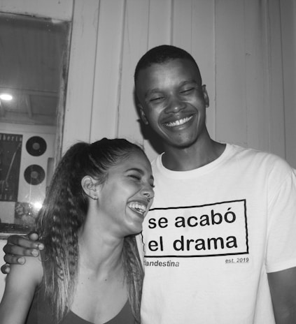 A black and white photograph of two people laughing, standing closely together. They appear to be enjoying a lighthearted moment. One person is wearing a T-shirt with Spanish text.