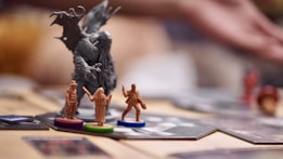 A close-up view of a board game setup featuring small, detailed figurines. In the foreground, there are three color-coded miniature figures on round bases, each representing a different character. Behind them stands a larger, ominous figure resembling a monster or creature, with outstretched claws. The background is softly blurred, focusing attention on the figures.