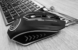 A sleek, futuristic-looking computer mouse with intricate design patterns sits on a wooden desk next to a keyboard. The mouse features smooth lines and geometric shapes, and the keyboard keys are clearly visible.